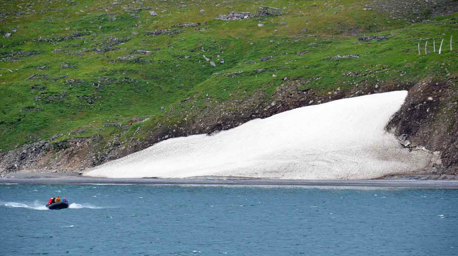 A small inflatable boat with passengers moving across blue water near a green hillside with a patch of snow