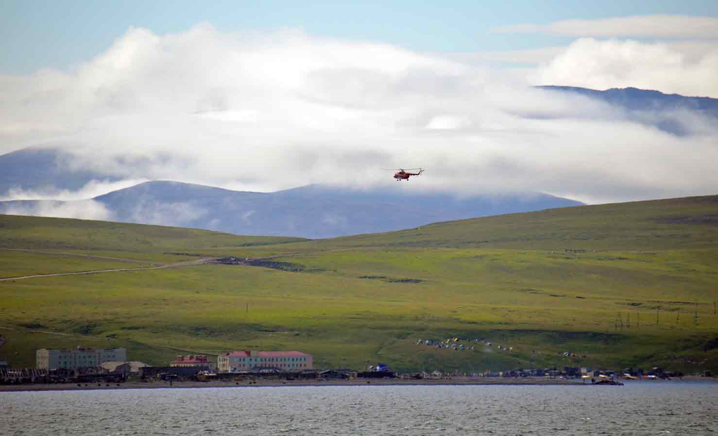 A helicopter flying low over a coastal town with colorful buildings, green hills and cloudy mountains in the background