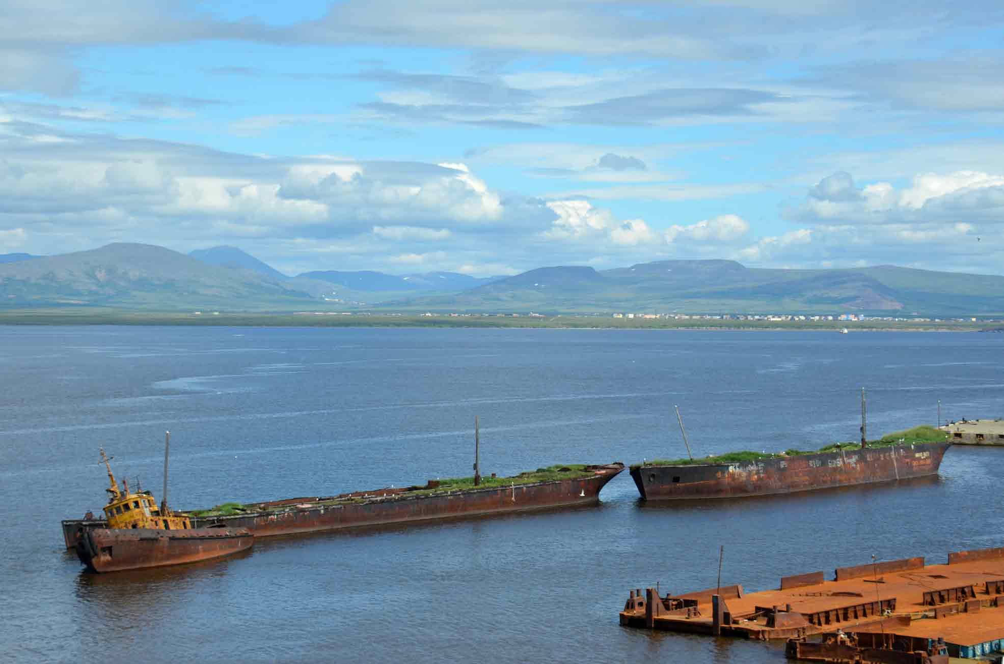 Rusted shipwrecks and a barge in a wide bay with mountains in the background under a partly cloudy sky