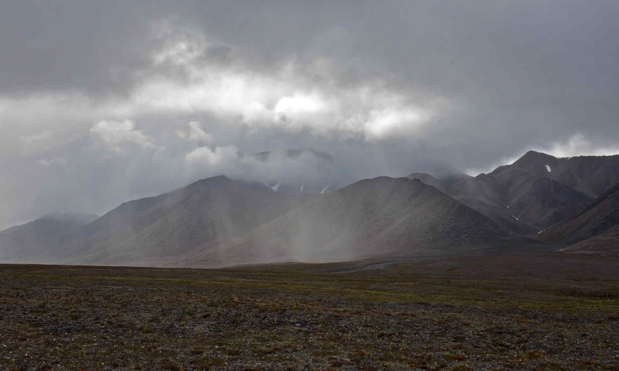 Wide barren mountain landscape under a heavy grey stormy sky with rain visible over distant peaks; muted brown and grey tones