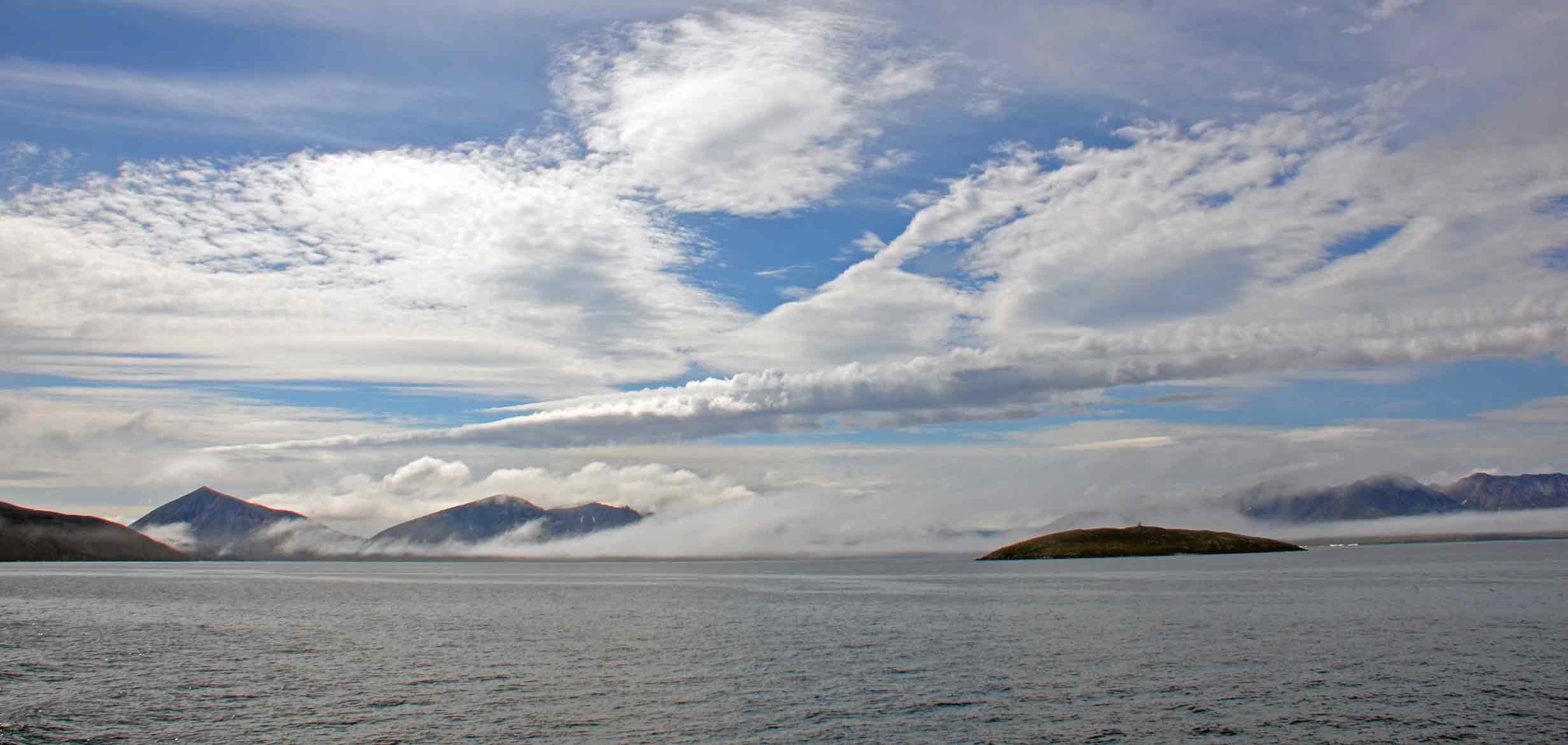 Wide seascape with layered clouds over distant misty mountains and a small island in calm water; blue, white and grey palette